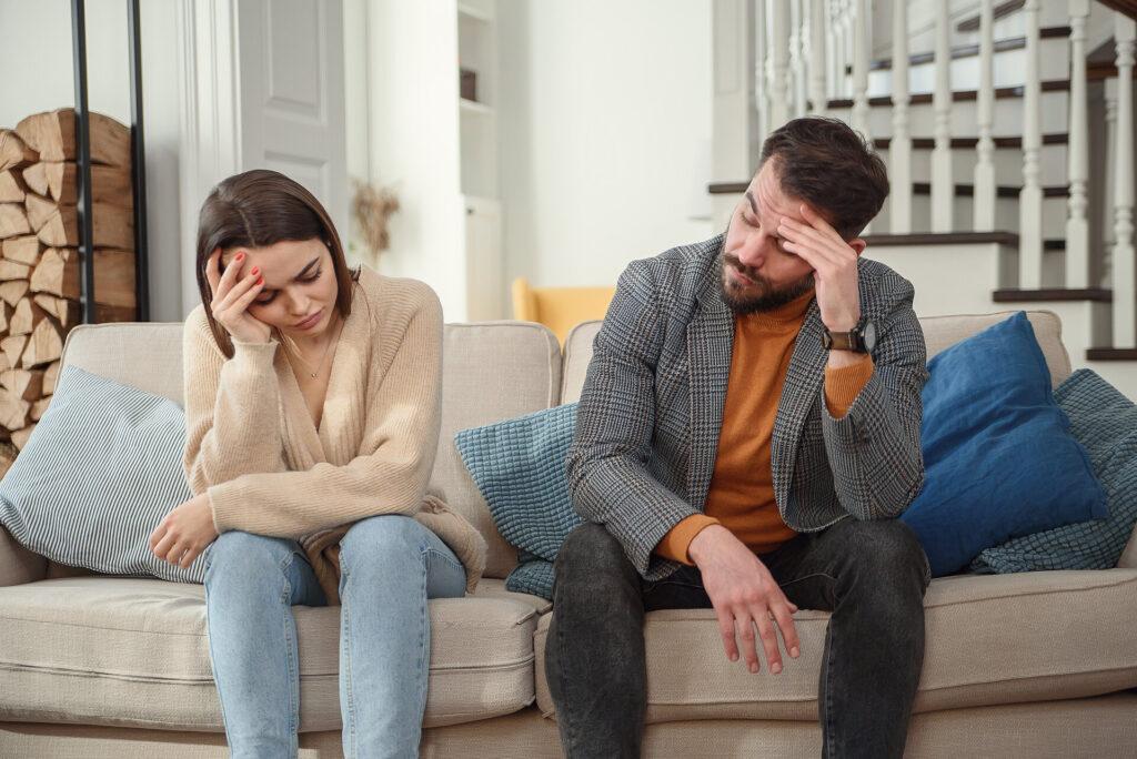 Unglückliches Paar, das nebeneinander traurig auf der Couch sitzt, nach unten schaut und sich voneinander abwendet.